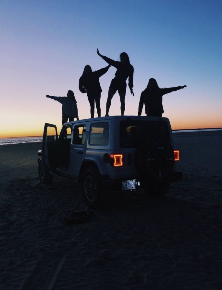 four people standing on the back of a jeep with their arms in the air at sunset