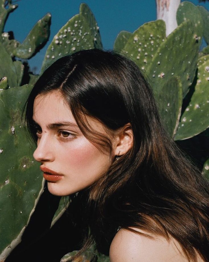 a woman with long hair standing next to a green plant and looking off into the distance