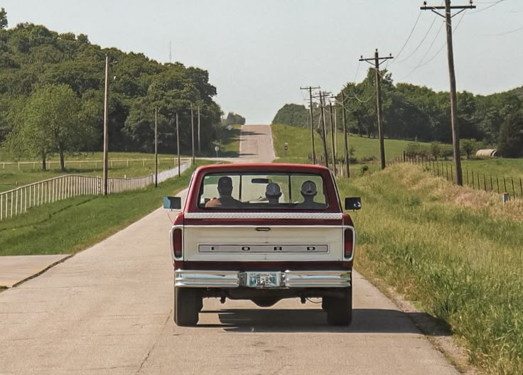 a red pickup truck driving down a rural road with two people in the back seat