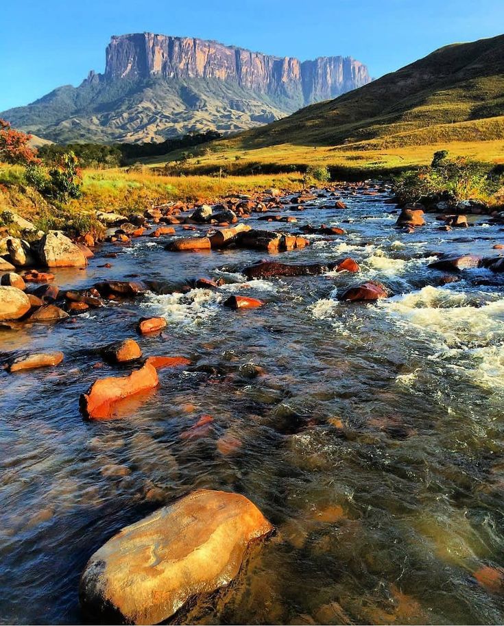 a river running through a lush green valley surrounded by mountains in the distance with rocks and grass on both sides