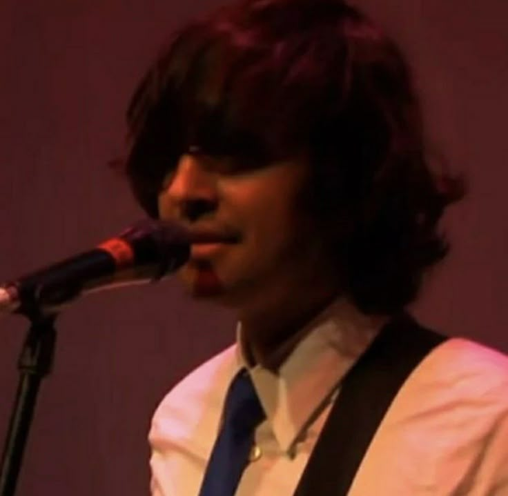 a young man with dark hair wearing a white shirt and blue tie singing into a microphone