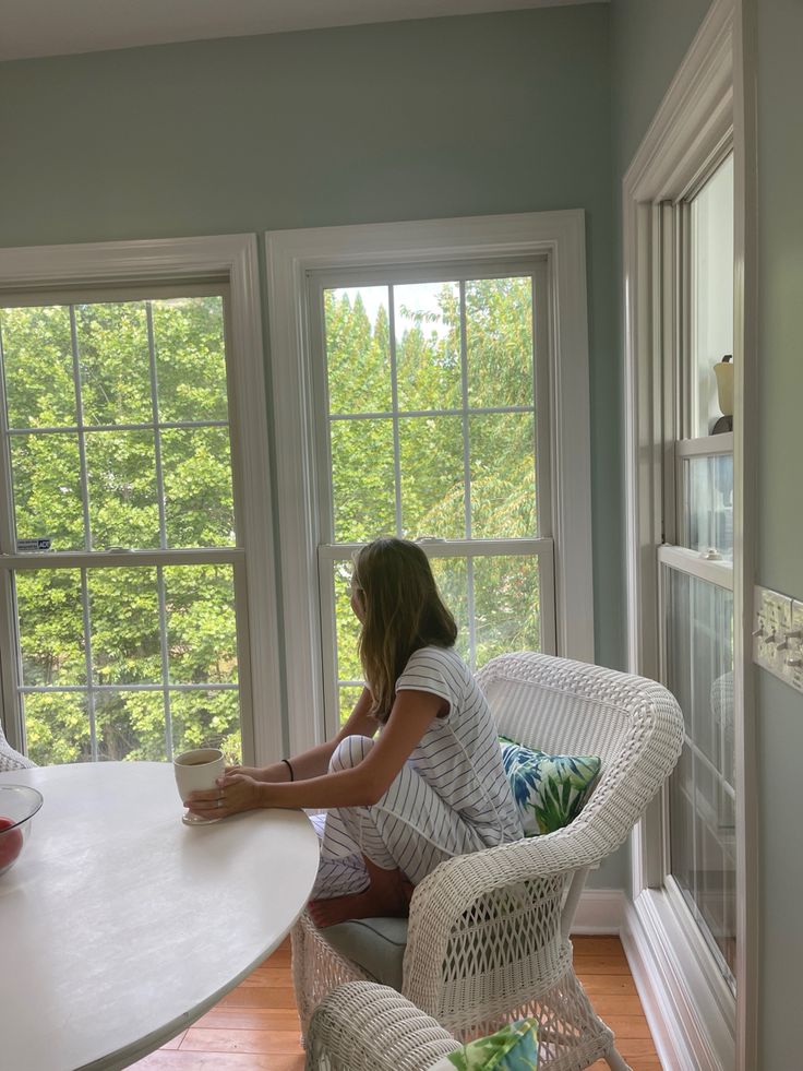 a woman sitting at a table in front of two large windows looking out onto the woods