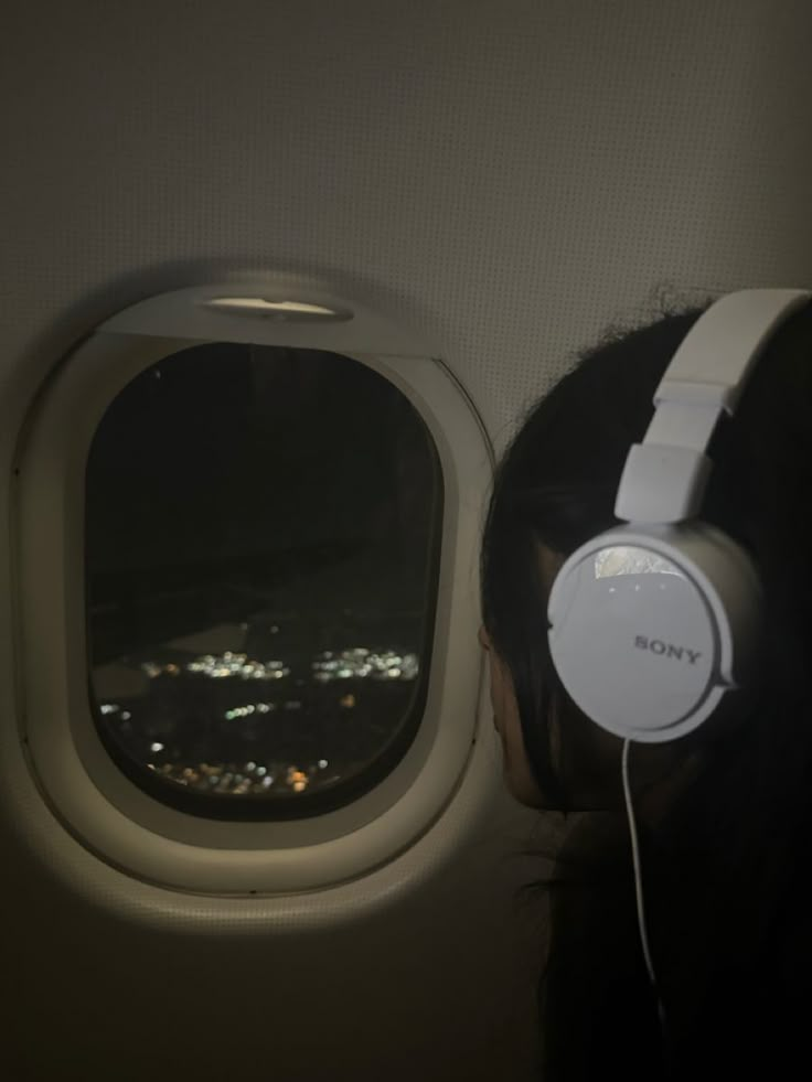 a woman with headphones on looking out an airplane window at the city lights below