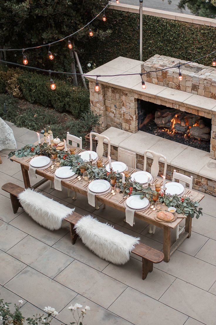 an outdoor dining table set up with white plates and place settings for dinner, next to a fireplace