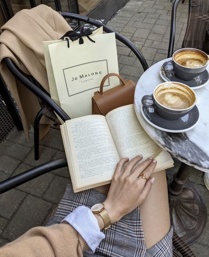 a woman is reading a book and drinking coffee while sitting at an outdoor table with two cups of coffee in front of her