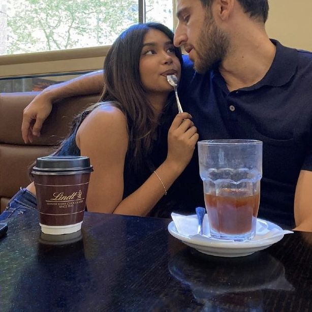 a man and woman sitting at a table with drinks in front of them, one holding a spoon to the other's mouth