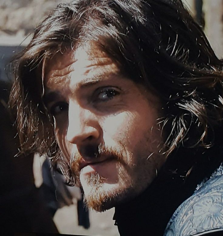 a man with long hair and beard standing in front of a car looking at the camera