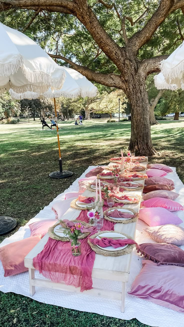 a long table covered in pink and white pillows under an umbrella next to a tree