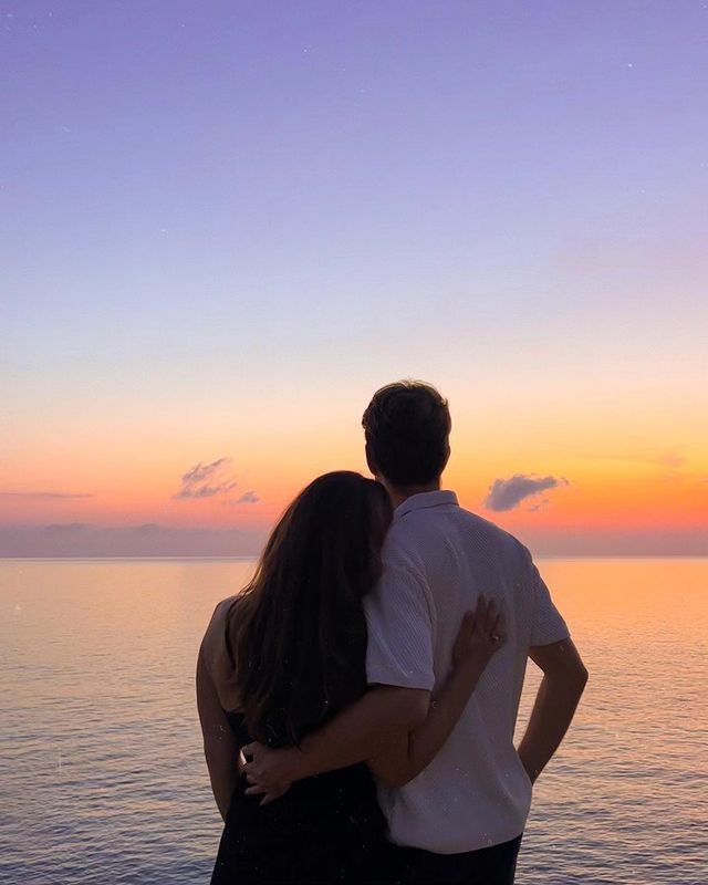 a man and woman standing next to each other near the ocean at sunset or sunrise