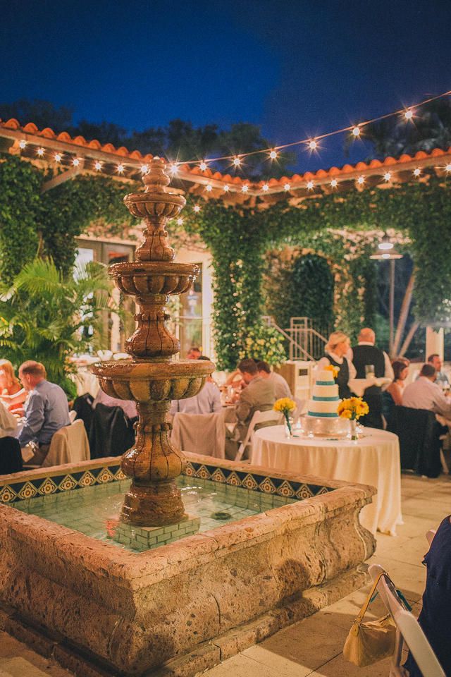 people sitting at tables in front of a fountain with lights strung over it and greenery on the walls