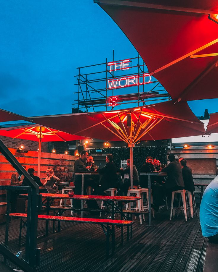 people are sitting at tables under umbrellas on the roof top bar in front of an illuminated sign that says the world is