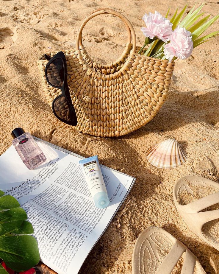 an open book on the sand next to a basket with flowers in it and some flip flops