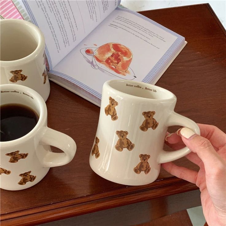 two coffee mugs sitting on top of a wooden table next to an open book