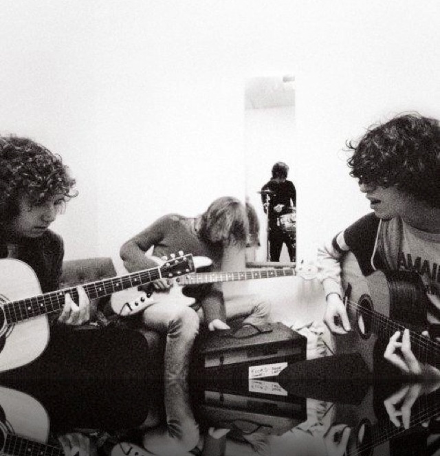 three young men sitting in a living room playing guitars and singing into microphones, with the words inside the inside out kooks above them