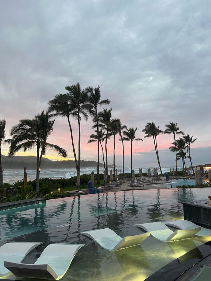 an outdoor swimming pool with lounge chairs and palm trees in the background at sunset or dawn