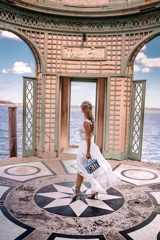 a woman in a white dress is walking through an ornate building by the ocean with her handbag