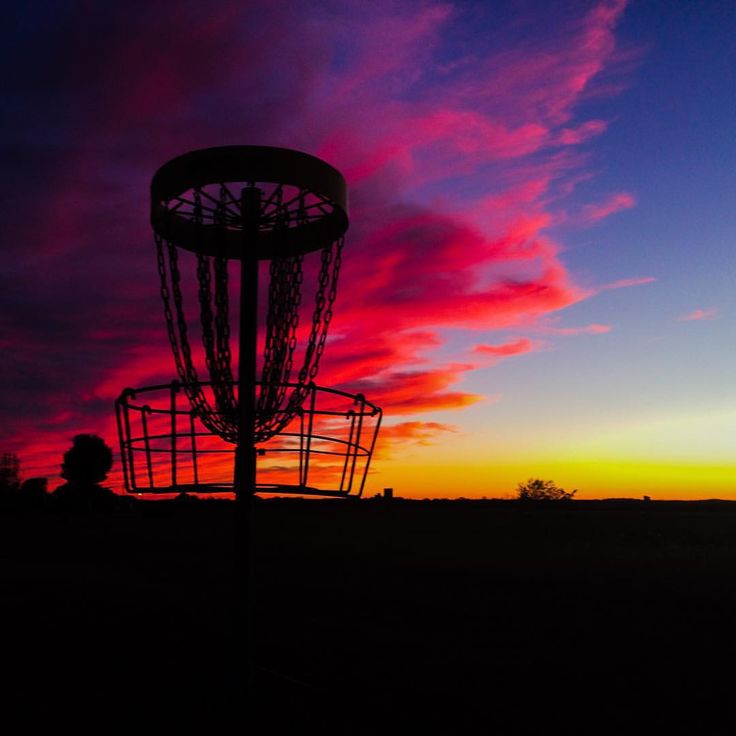 the silhouette of a frisbee golf cage against a colorful sky with clouds in the background