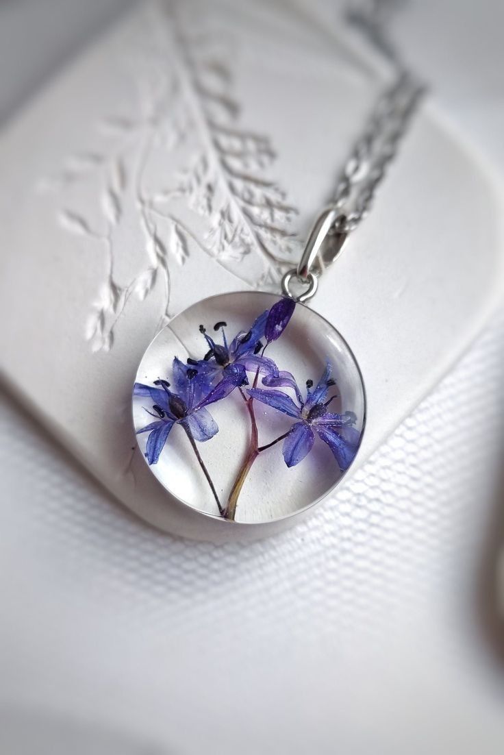 a glass pendant with purple flowers in it sitting on a white card and silver chain