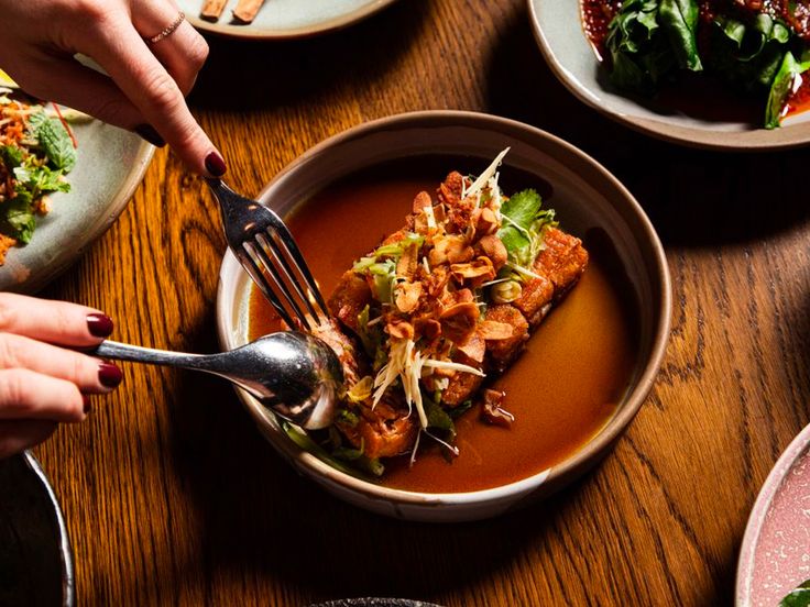 several plates of food on a wooden table with utensils in each bowl and hands holding forks