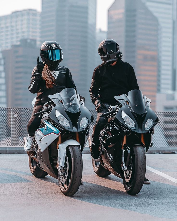 two people wearing helmets are riding motorcycles in the parking lot with tall buildings behind them