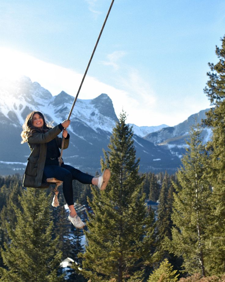 a woman is swinging on a rope in the air with mountains and trees behind her