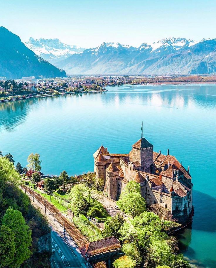 an old castle sits on the shore of a lake with mountains in the background and blue water