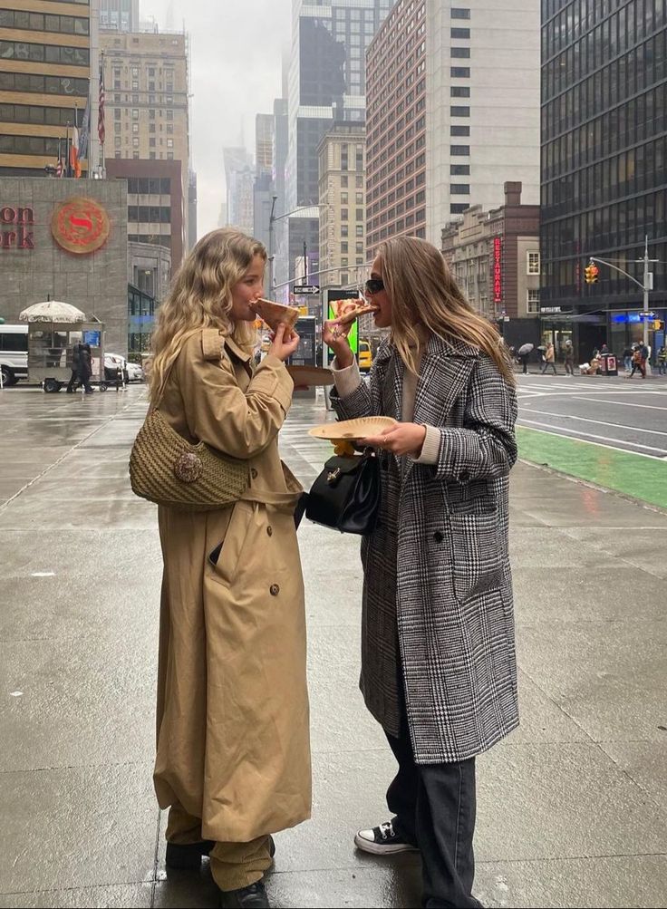 two women standing on the side of a road talking to each other and eating food