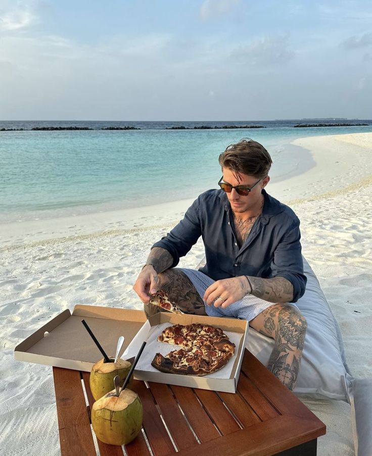 a man sitting at a table with pizza and an apple on the beach in front of him