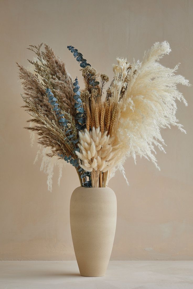 a white vase filled with dried flowers and grass in front of a beige wall background