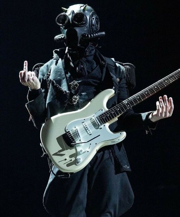 a man in a gas mask holding a guitar and wearing a black outfit with his mouth open