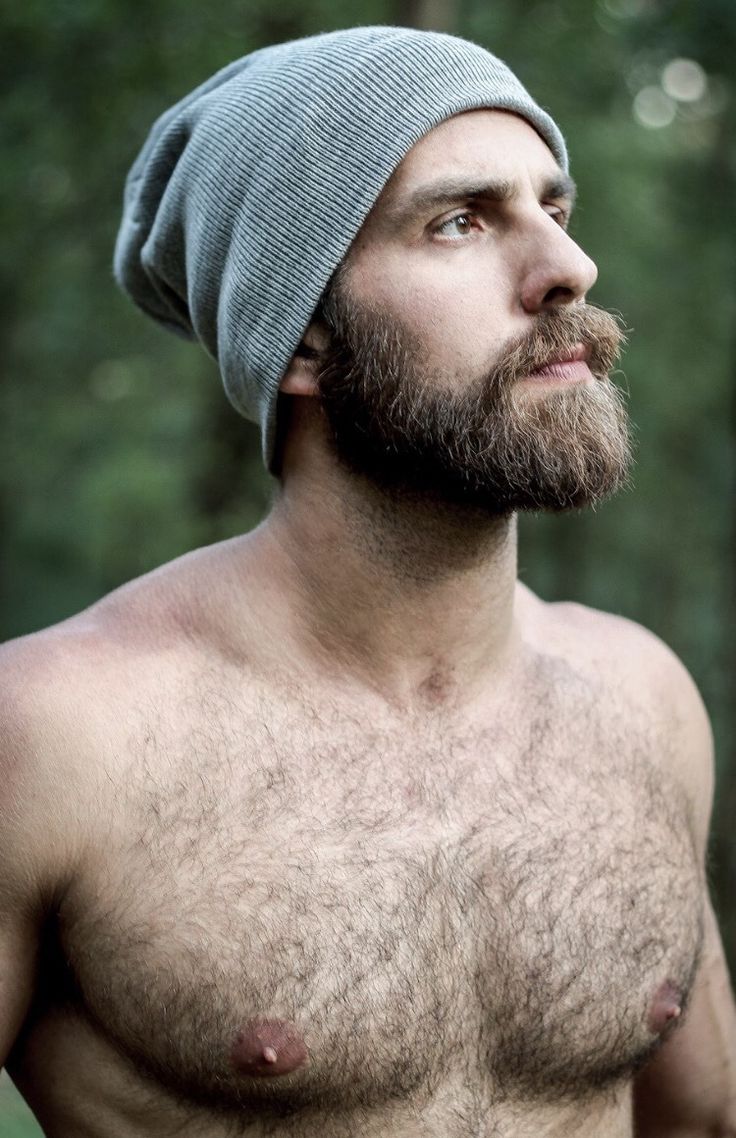 a shirtless man wearing a beanie looks up into the sky while standing in front of trees