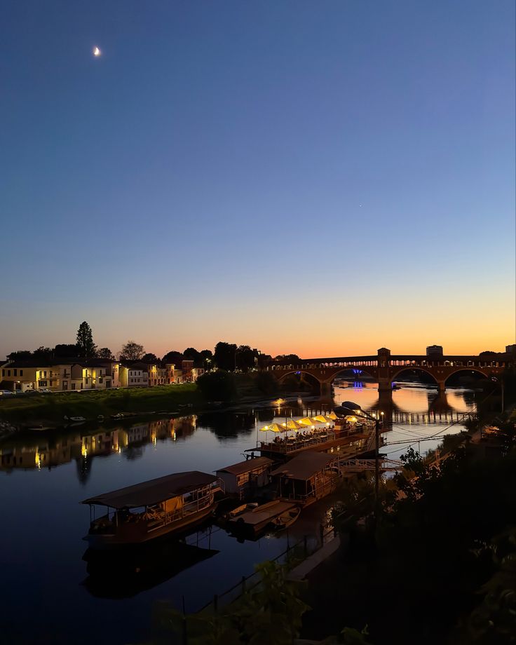 boats are docked in the water at dusk near a bridge and buildings with lights on them