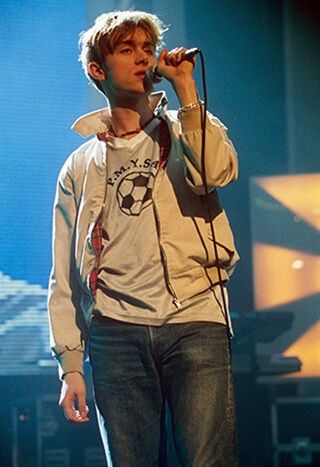 a male in a white shirt is singing into a microphone and holding a microphone up to his ear