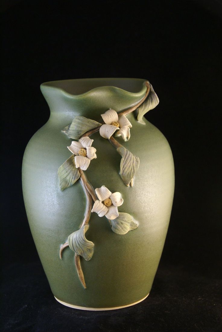 a green vase with white flowers and leaves on the outside, against a black background