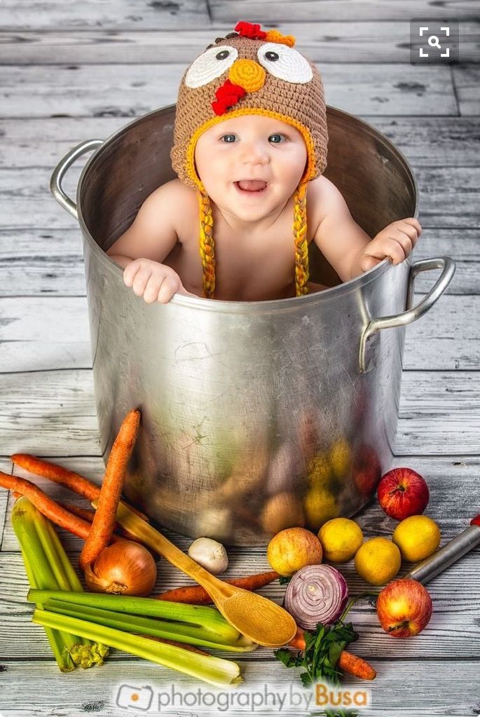 a baby in a metal bucket with vegetables and fruits around it, wearing a turkey hat