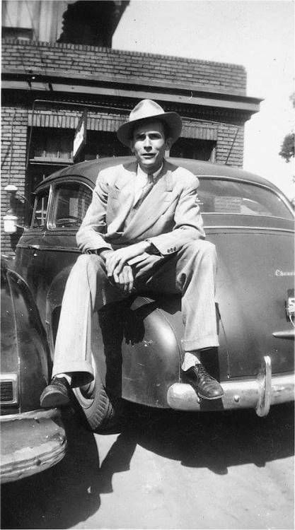 a man in a suit and hat sitting on the hood of an old fashioned car