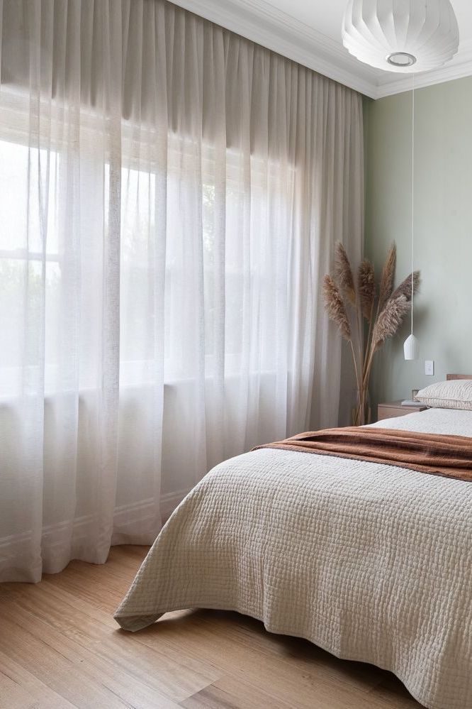 a bed sitting next to a window covered in white drapes and pillows on top of a hard wood floor