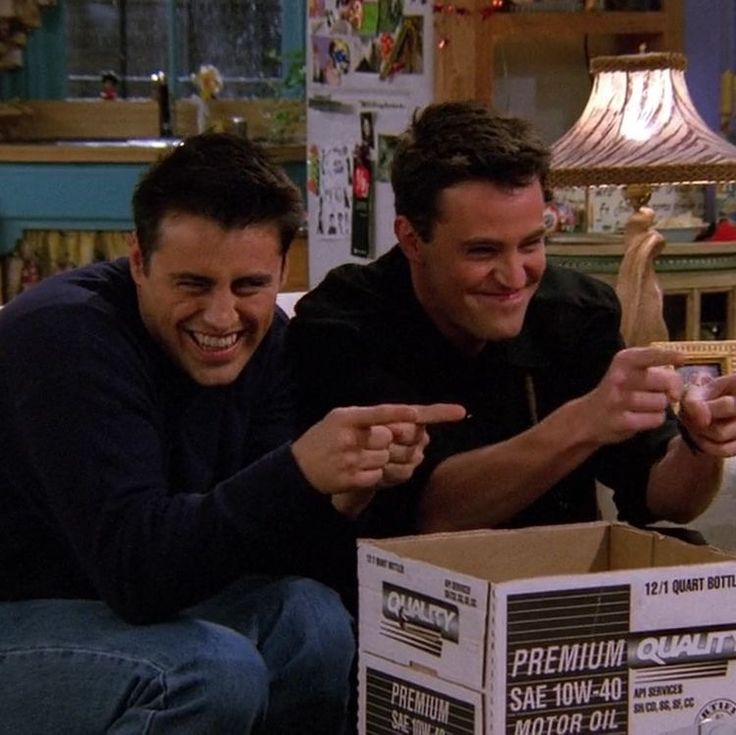 two men sitting next to each other on the floor pointing at something in a box