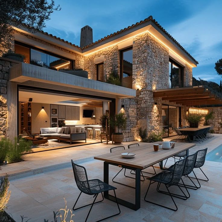 an outdoor dining area next to a swimming pool at night with lights on the house