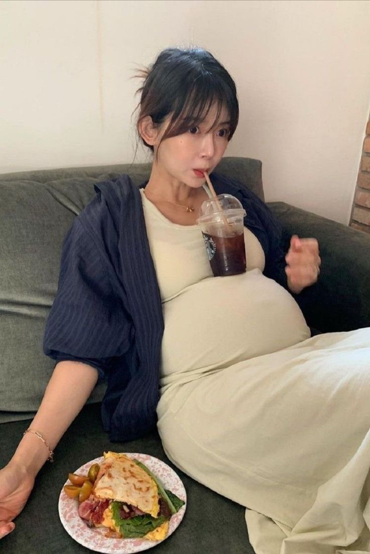 a pregnant woman sitting on a couch drinking from a straw in her mouth while holding a plate of food