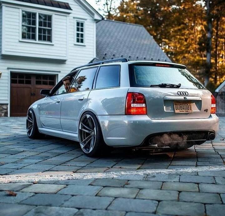 the rear end of a silver car parked in front of a white house on a cobblestone driveway