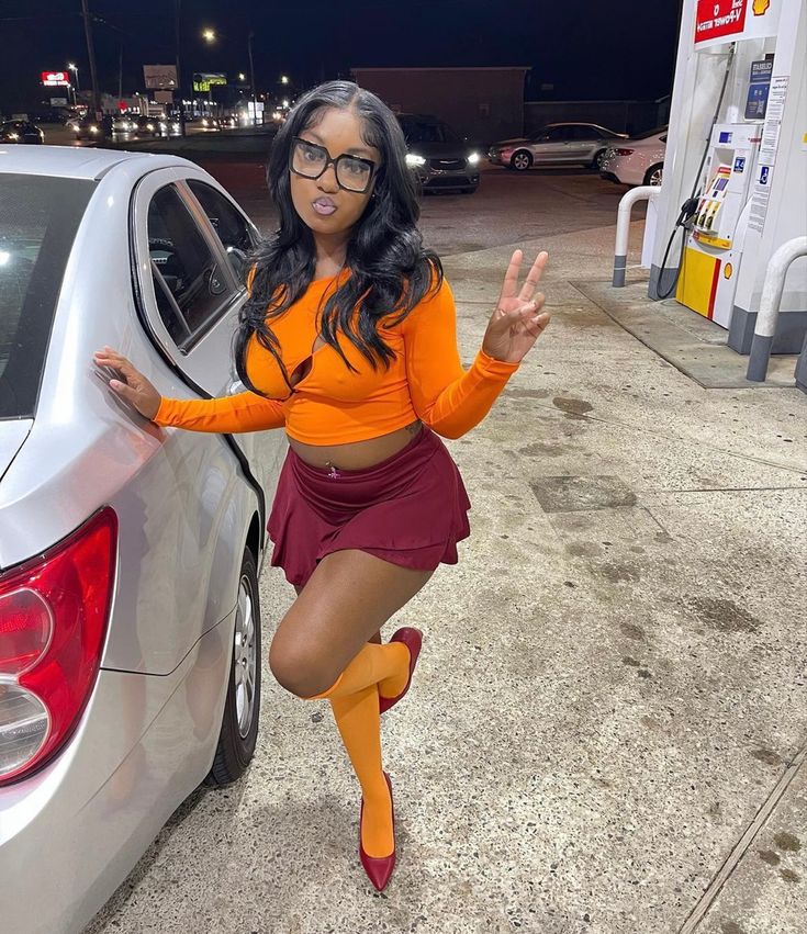 a woman in an orange top and maroon skirt is leaning against a white car at a gas station