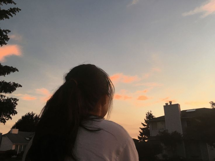 a woman with long hair standing in front of the sun at dusk, looking up into the sky