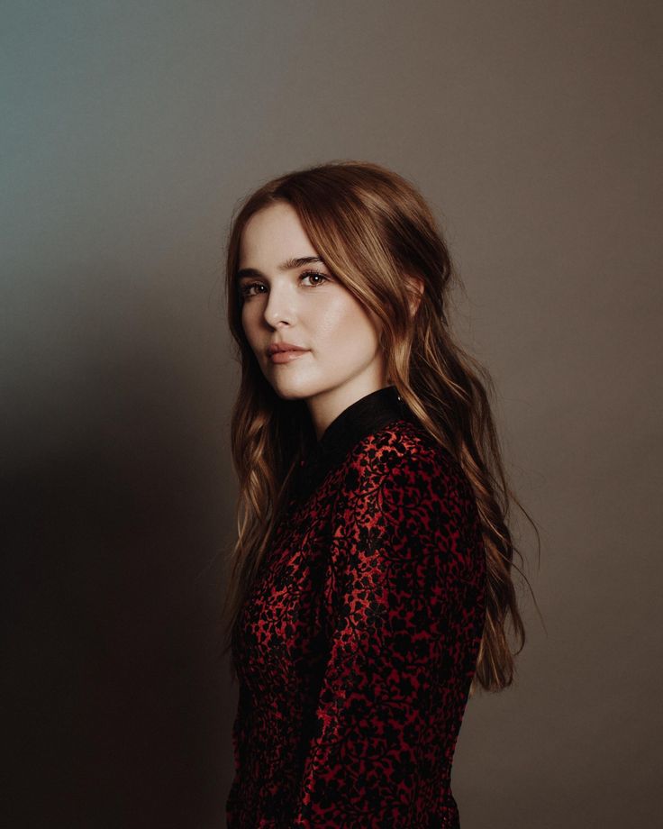 a woman with long hair wearing a red and black top posing for a photo in front of a gray wall