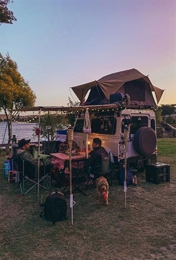 a camper van parked next to a lake with people sitting in the shade under it