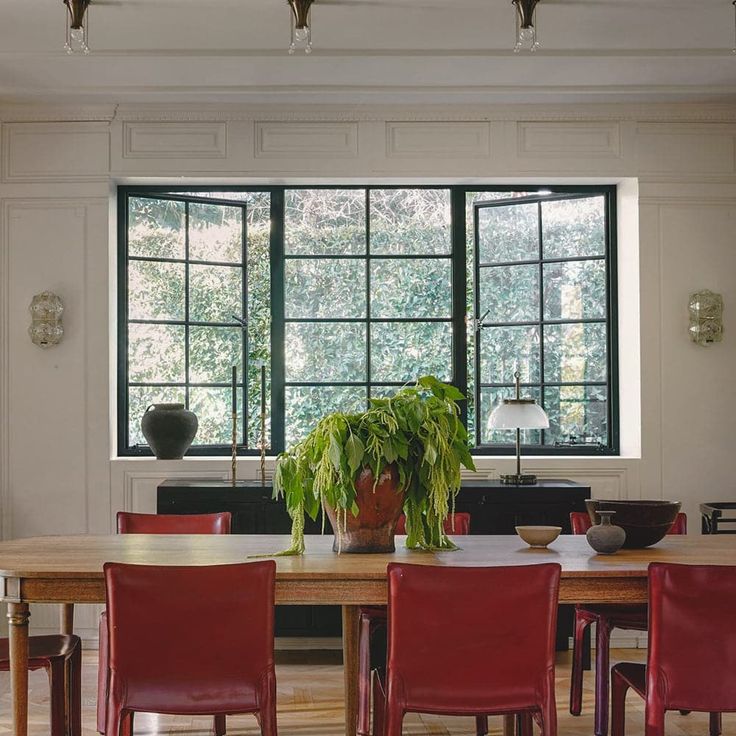 a dining room table with red chairs and a potted plant on the table in front of two large windows