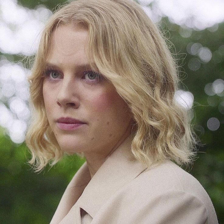 a woman with blonde hair wearing a beige coat and looking off into the distance while standing in front of trees