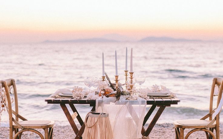 a table set up on the beach with candles and flowers in vases, along with two chairs