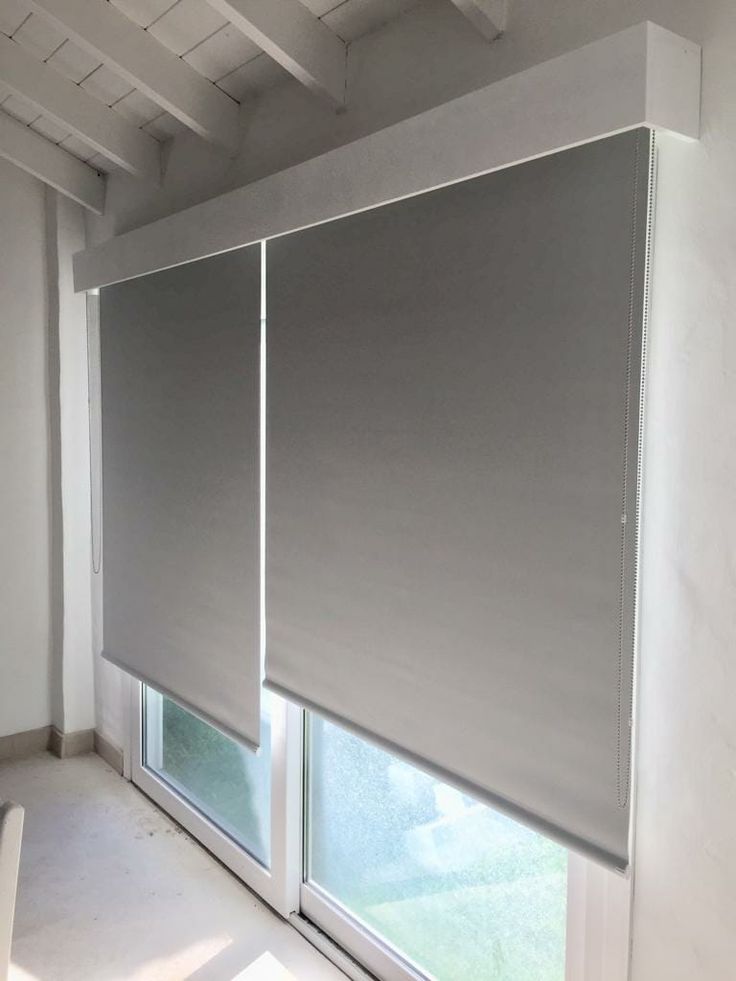 an empty room with two windows covered in gray roller shades and a white chair next to the window