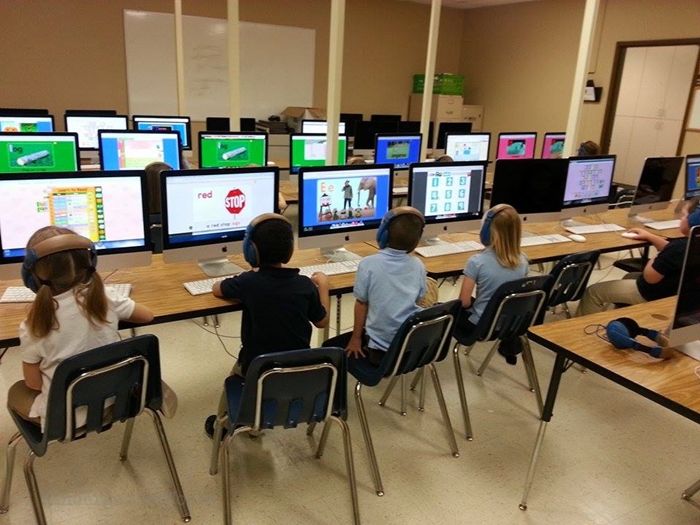 children are sitting at desks with computer screens on the wall and in front of them